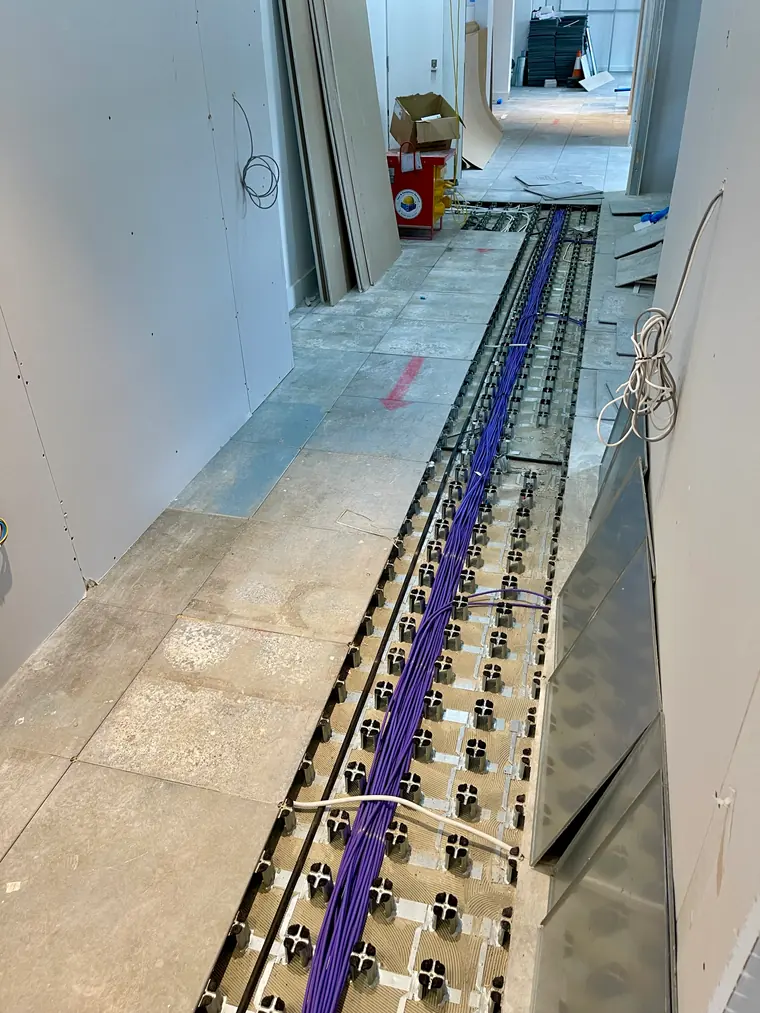 A dressed loom of violet Cat6 cabling running along the void of a raised access floor during an office fit-out, floor tiles lifted along the route