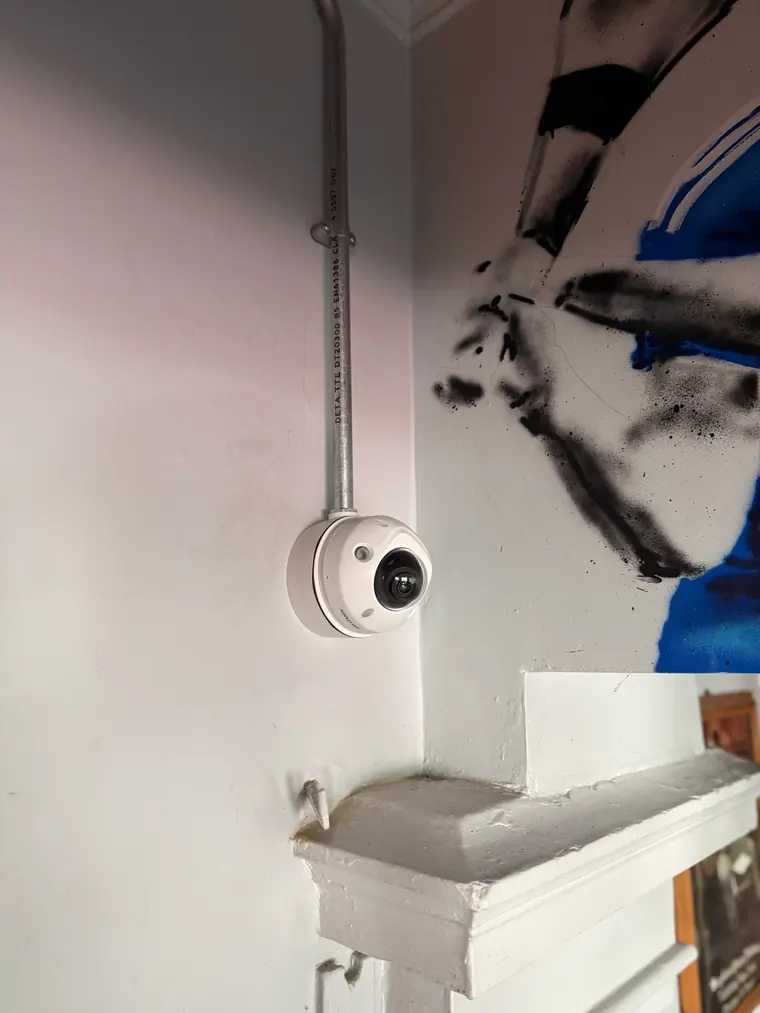 A compact white dome camera mounted inside on galvanised steel conduit, against a wall with a painted mural