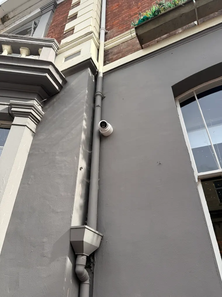 A white turret camera mounted high on the rendered facade of a period building, positioned beside the rainwater pipe to cover the frontage
