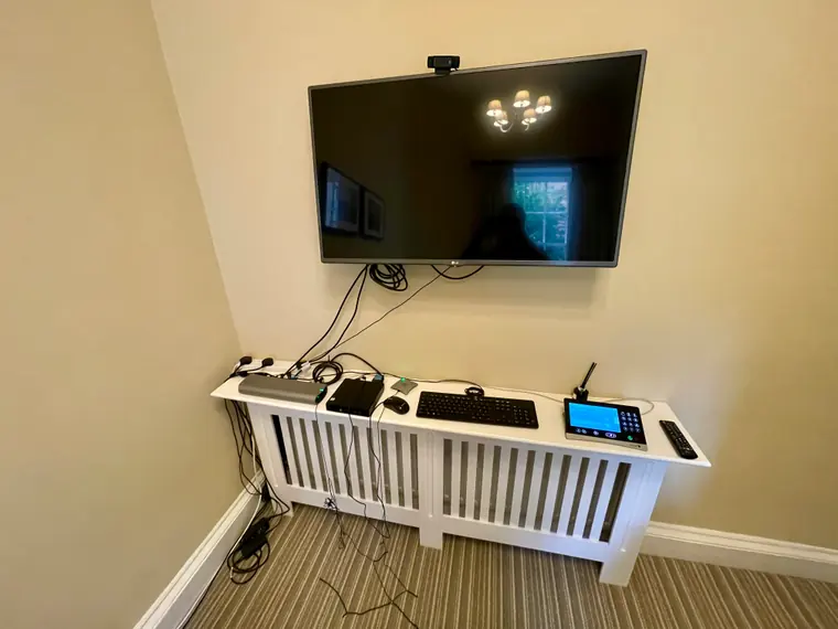 Before: a wall-mounted TV with a consumer webcam balanced on top, a keyboard and mouse resting on a radiator cover, and loose cables trailing down the wall